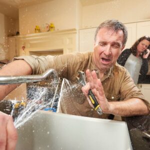 man getting sprayed by faucet. woman on phone in the background